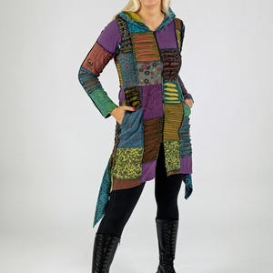 May include: A patchwork hooded jacket in shades of purple, green, and brown. The jacket features a variety of patterned fabric squares, a hood, and pockets. The person is wearing black leggings and black knee-high boots.