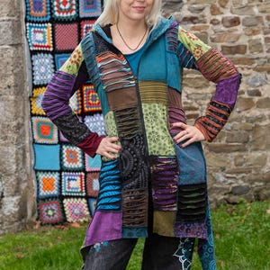 May include: A multi-coloured patchwork jacket with a hood and zip closure. The jacket features a mix of patterns and textures, including teal, purple, and brown. The person is wearing dark trousers with a floral pattern. A colourful crocheted wall hanging is in the background.