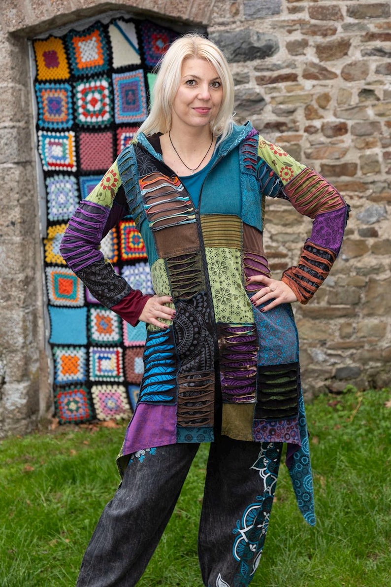 May include: A multi-coloured patchwork jacket with a hood and zip closure. The jacket features a mix of patterns and textures, including teal, purple, and brown. The person is wearing dark trousers with a floral pattern. A colourful crocheted wall hanging is in the background.