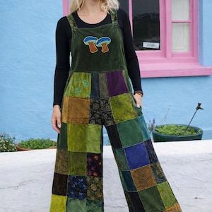 Może przedstawiać: Zielone ogrodniczki w stylu patchwork z motywem grzyba na piersi. Szerokie nogawki są noszone na czarnej bluzce z długim rękawem. Patchwork zawiera różne odcienie zieleni, brązu i niebieskiego.