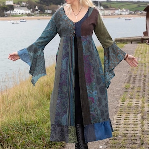 Puede incluir: Un abrigo largo de patchwork en tonos turquesa, azul, verde y marrón. El abrigo largo tiene mangas acampanadas y una botonadura frontal. Se lleva con pantalones y botas negras. El fondo muestra una masa de agua y un cielo nublado.