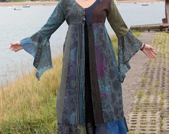 May include: A patchwork duster coat in shades of teal, blue, green, and brown. The long coat features bell sleeves and a button-down front. The coat is worn with black pants and boots. The background shows a body of water and a cloudy sky.