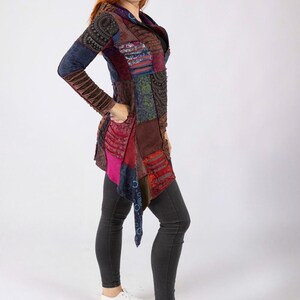 May include: A multi-coloured patchwork jacket with long sleeves and a draped front. The jacket features shades of blue, brown, red, and pink. The model is wearing dark grey trousers and white trainers.