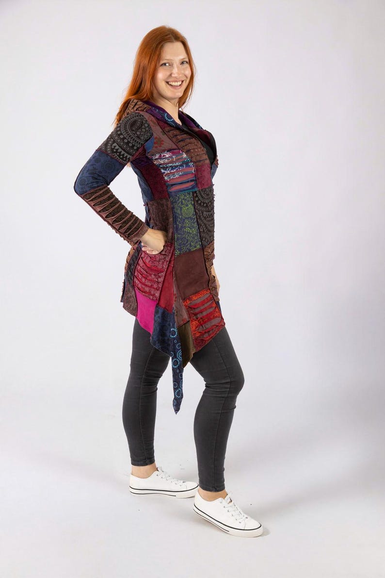 May include: A multi-coloured patchwork jacket with long sleeves and a draped front. The jacket features shades of blue, brown, red, and pink. The model is wearing dark grey trousers and white trainers.