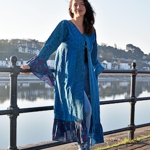 Puede incluir: Un abrigo largo y fluido de color azul con cuello en V y mangas acampanadas. El abrigo presenta un diseño estampado y un dobladillo con volantes. Las mangas tienen un estampado floral morado y azul. El abrigo se lleva sobre leggings azules y zapatillas blancas.