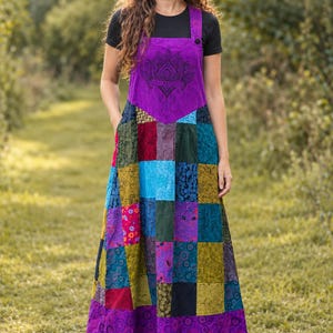 May include: A floor-length patchwork pinafore dress featuring a variety of colourful fabric squares in shades of purple, blue, green, and red. The bodice displays a black lotus flower print, and the skirt includes side pockets and black boots.