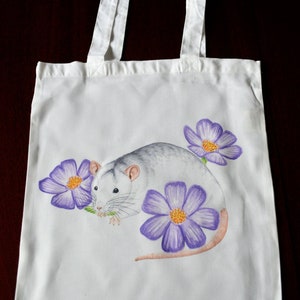 May include: White canvas tote bag with a hand-painted design of a gray rat with purple flowers. The rat is sitting on a flower and eating a green leaf.