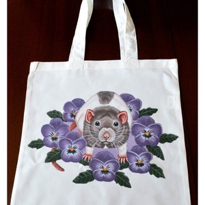 May include: White tote bag with a colourful illustration of a rat surrounded by purple pansies. The rat has grey and white fur, with a pink nose and ears. The tote bag has white handles.