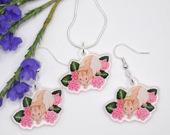 May include: A set of jewelry featuring a rat with pink flowers. The necklace and earrings are silver chain with a rat charm. The rat is white with pink eyes and is surrounded by pink flowers.