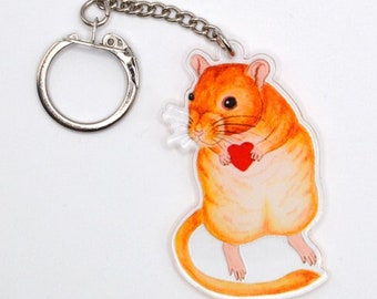 May include: A keychain featuring a cartoon illustration of a gerbil holding a red heart. The gerbil is orange and white and has a pink nose. The keychain is made of clear acrylic.