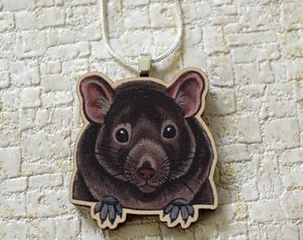 May include: A wooden pendant necklace featuring a black and brown rat with a white nose and whiskers. The rat is looking directly at the viewer with its paws resting on the edge of the pendant.