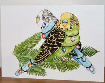 May include: A watercolor greeting card featuring two parakeets perched on a sprig of evergreen. The birds are adorned with colorful ornaments. One parakeet has black and yellow feathers, while the other is green and yellow. The card is on a wooden surface.