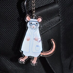 May include: A white mouse wearing a lab coat and goggles is attached to a silver keyring. The keyring is attached to a black bag.