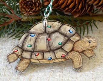 May include: A hand-painted Christmas ornament featuring a turtle with a shell decorated with colorful Christmas lights. The ornament is made of clear acrylic and has a string for hanging.