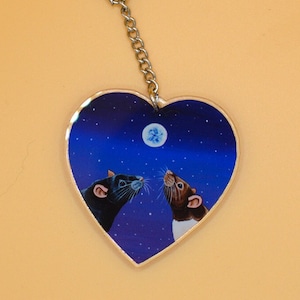 May include: A heart-shaped keyring with a night sky design featuring two rats looking at a moon. The rats are black and white and the sky is a dark blue with white stars.