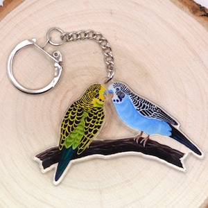 May include: A silver keychain featuring two parakeets, one blue and one green, perched on a branch. The parakeets are facing each other and appear to be kissing.