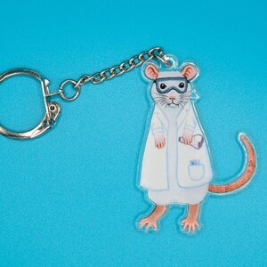 May include: A white lab coat keychain featuring a cartoon mouse wearing goggles and holding a test tube. The mouse has a brown tail and pink ears.