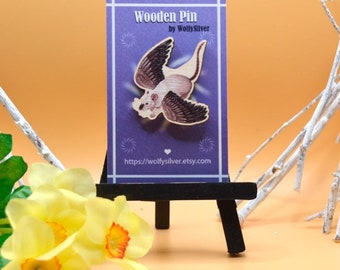 May include: A wooden pin featuring a white and gray flying creature with wings. The pin is on a purple background with the text "Wooden Pin by WolfySilver" and a website address.