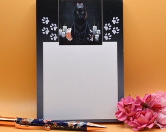 May include: A black notepad with a black cat and candles illustration. The notepad has a white writing surface and a black border. The illustration features a black cat sitting on a pedestal with candles on either side. The cat has a crown on its head and is looking at the viewer. The notepad is on a light orange surface.