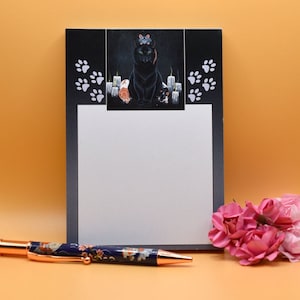 May include: A black notepad with a black cat and candles illustration. The notepad has a white writing surface and a black border. The illustration features a black cat sitting on a pedestal with candles on either side. The cat has a crown on its head and is looking at the viewer. The notepad is on a light orange surface.