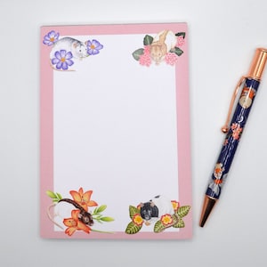 May include: Pink notepad with a white interior and a pink border. The notepad has floral illustrations with mice on each corner. The notepad is next to a pen with a floral design.