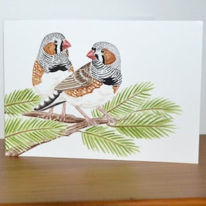 May include: A watercolour greeting card showing two zebra finches on a branch with green leaves. The birds have grey and white striped heads, brown wings, and red beaks. The card is on a wooden surface.