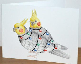 May include: A folded greeting card featuring a watercolor illustration of two cockatiels adorned with colorful Christmas lights. The birds have yellow crests and red cheek patches. The card is white and sits on a wooden surface.