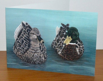 May include: A greeting card featuring a painting of two ducks swimming in a blue pond. The duck on the left is brown and gray, while the duck on the right is brown with a green head.