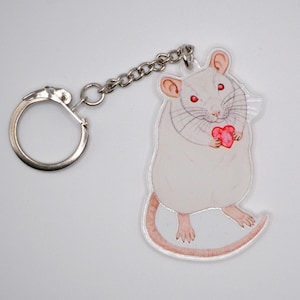May include: White rat keyring with a red heart. The rat is holding the heart in its paws. The keyring is made of clear acrylic.