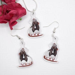 May include: A set of silver rat earrings and a necklace. The rat charms are white with black markings and have a star on their chest. The earrings and necklace are on a white background.