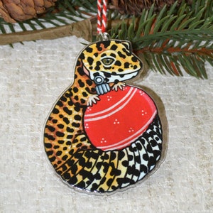 Peut inclure: Une décoration de Noël représentant un gecko léopard tenant une boule rouge. Le gecko est illustré dans un style cartoon avec des taches noires et jaunes. La décoration est en bois et possède une ficelle pour l'accrocher.