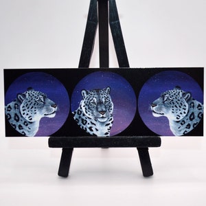 Puede incluir: Tres pinturas de leopardo de las nieves sobre un fondo negro. Cada pintura presenta un leopardo de las nieves con una pose de cabeza diferente contra un cielo estrellado morado y azul.