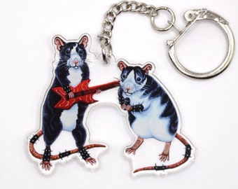 May include: A black and white rat playing a red electric guitar and a black and white rat holding a microphone. Both rats are wearing studded bracelets and have spiky tails. The rats are attached to a silver key chain.