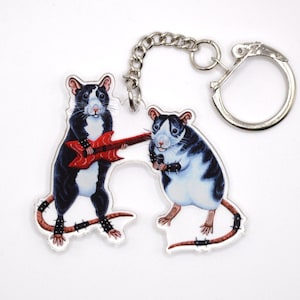 May include: A black and white rat playing a red electric guitar and a black and white rat holding a microphone. Both rats are wearing studded bracelets and have spiky tails. The rats are attached to a silver key chain.
