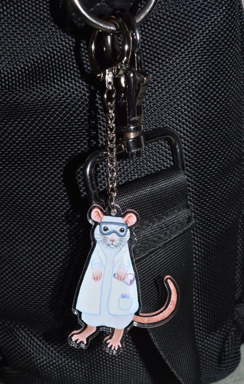 May include: A white mouse wearing a lab coat and goggles is attached to a silver keyring. The keyring is attached to a black bag.