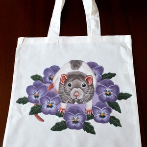 May include: White tote bag with a detailed illustration of a rat surrounded by purple pansies. The rat has grey and white fur, with pink ears and a focused gaze. The bag has white handles.