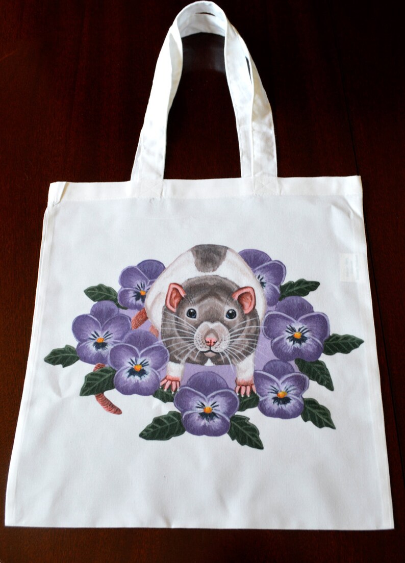May include: White tote bag with a detailed illustration of a rat surrounded by purple pansies. The rat has grey and white fur, with pink ears and a focused gaze. The bag has white handles.