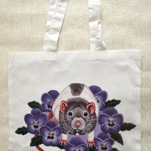 May include: White tote bag with a rat illustration surrounded by purple pansies. The rat has a black and white coat and is centered on the bag. White handles extend from the top, made of a sturdy fabric.