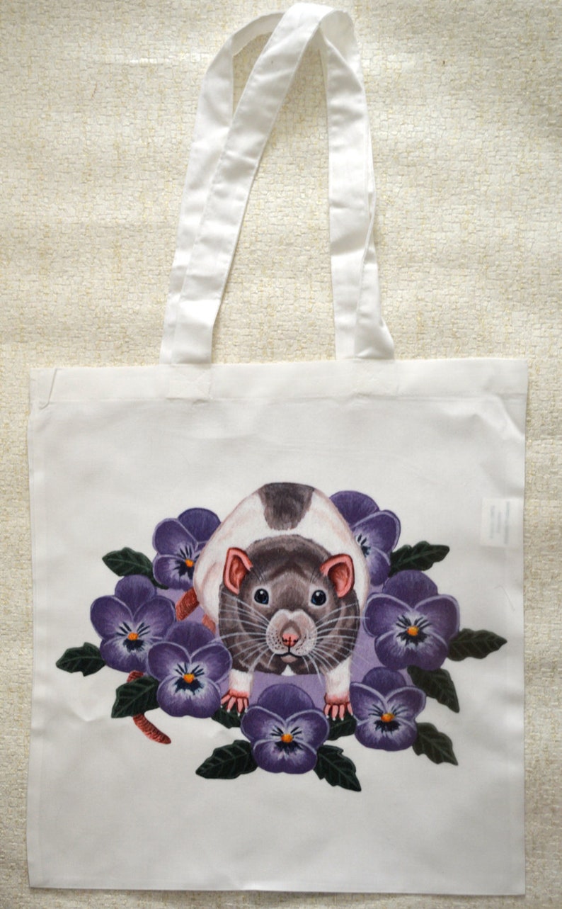 May include: White tote bag with a rat illustration surrounded by purple pansies. The rat has a black and white coat and is centered on the bag. White handles extend from the top, made of a sturdy fabric.