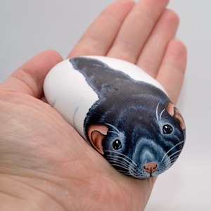 May include: Hand-painted rock art depicting a black rat with white whiskers and pink nose. The rat is painted in a realistic style with details on the fur and eyes.