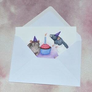 May include: A birthday card featuring two rats wearing party hats and looking at a cupcake with a lit candle. The rats are grey and white with pink noses and red eyes. The cupcake is pink with blue icing and a single blue candle.