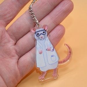 May include: A white lab rat wearing a white lab coat and goggles, holding a test tube. The rat is a keychain made of clear acrylic.