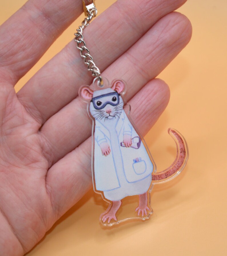 May include: A white lab rat wearing a white lab coat and goggles, holding a test tube. The rat is a keychain made of clear acrylic.