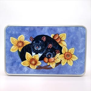 May include: A rectangular tin with a painted illustration of two Tasmanian devils nestled among yellow daffodils. The devils are black with white markings, set against a blue background with a silver border.