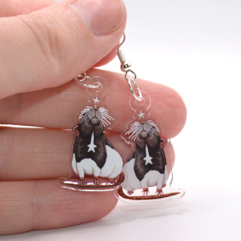 May include: A pair of clear acrylic earrings featuring a black and white rat with a star on its chest. The rat is looking up and has a star above its head. The earrings are on silver hooks.