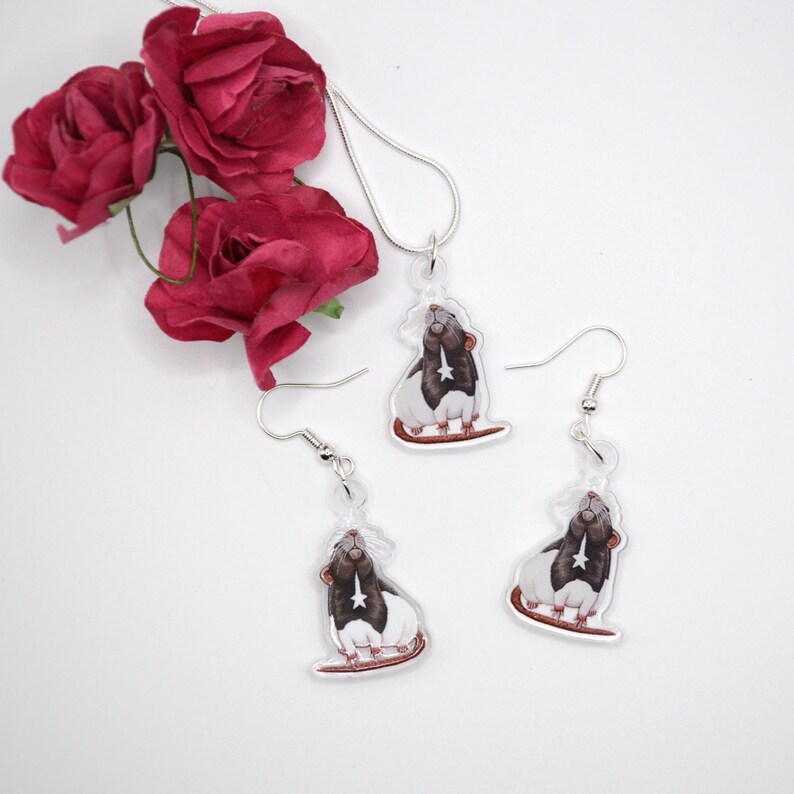 May include: A set of silver rat earrings and a necklace. The rat charms are white with black markings and have a star on their chest. The earrings and necklace are on a white background.