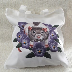 May include: White tote bag with a watercolour illustration of a rat surrounded by purple pansies. The rat has grey and white fur with pink ears and a pink nose. The tote bag has two white handles.