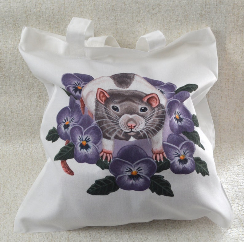 May include: White tote bag with a watercolour illustration of a rat surrounded by purple pansies. The rat has grey and white fur with pink ears and a pink nose. The tote bag has two white handles.