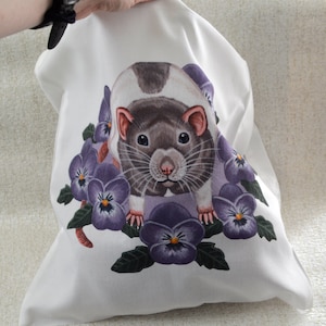 May include: White fabric bag with a detailed illustration of a rat and purple pansies. The rat has a brown and white coat, pink ears and nose. The pansies have yellow centres and green leaves.