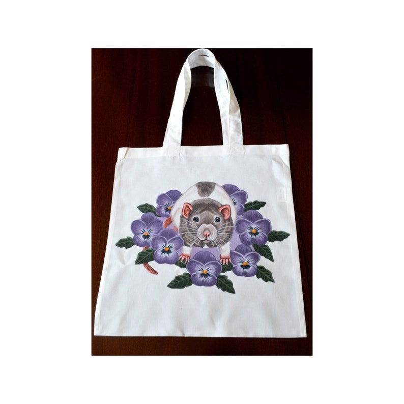 May include: White tote bag with a colourful illustration of a rat surrounded by purple pansies. The rat has grey and white fur, with a pink nose and ears. The tote bag has white handles.
